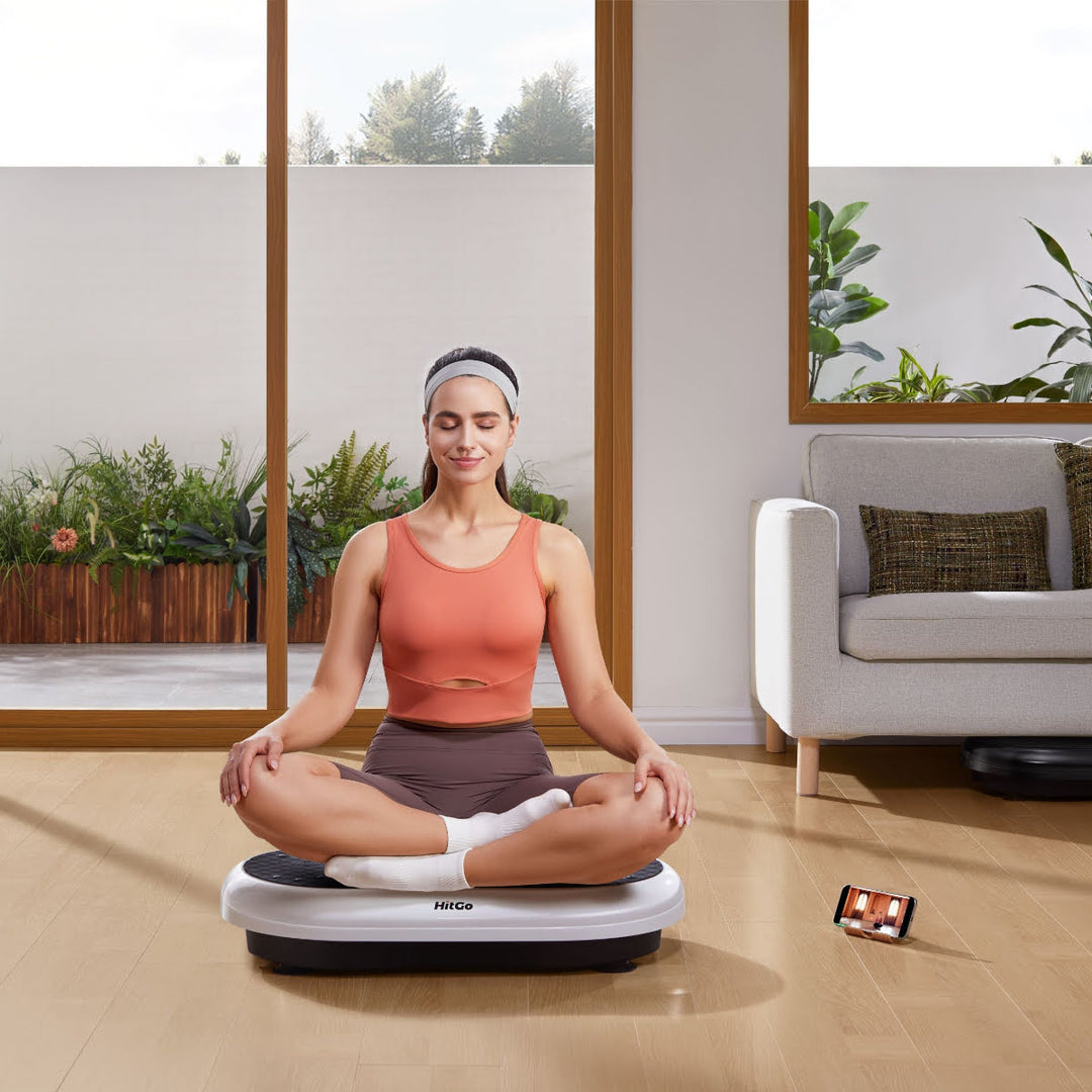 Woman sitting in the lotus position on vibration plate inside home. A phone is playing a yoga workout.