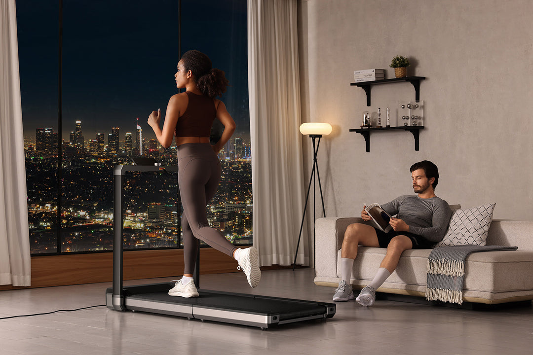 Woman exercising on a treadmill in a modern living room with a cityscape view, man reading on a couch.