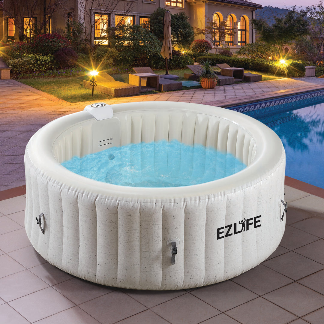 A white inflatable hot tub with the 'EZLife' brand logo, set against a beautiful sunset backdrop. In the background, there's a sparkling pool and a modern house, creating a serene and relaxing outdoor scene