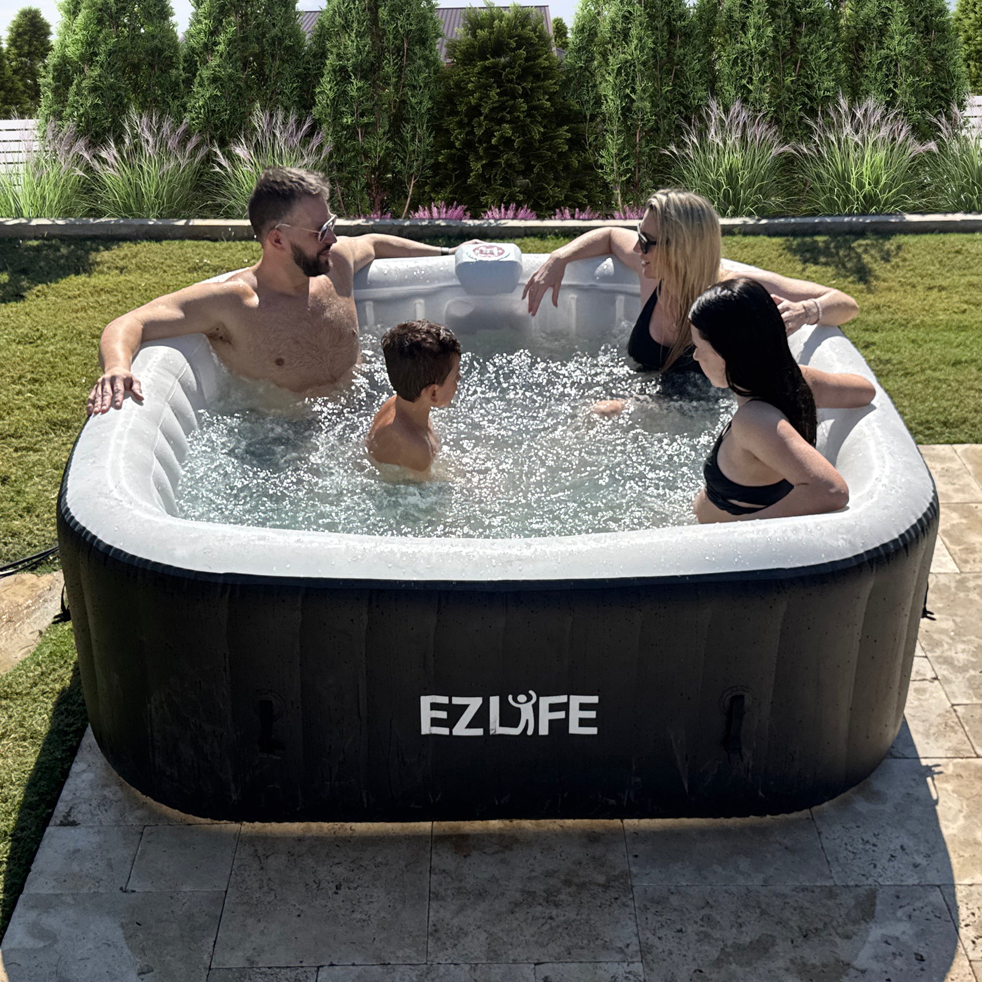 A black inflatable hot tub with the 'EZLife' brand logo. There are four people in the hot tub experiencing a nice soak under the sun in their backyard, with plants and flowers in the background. 