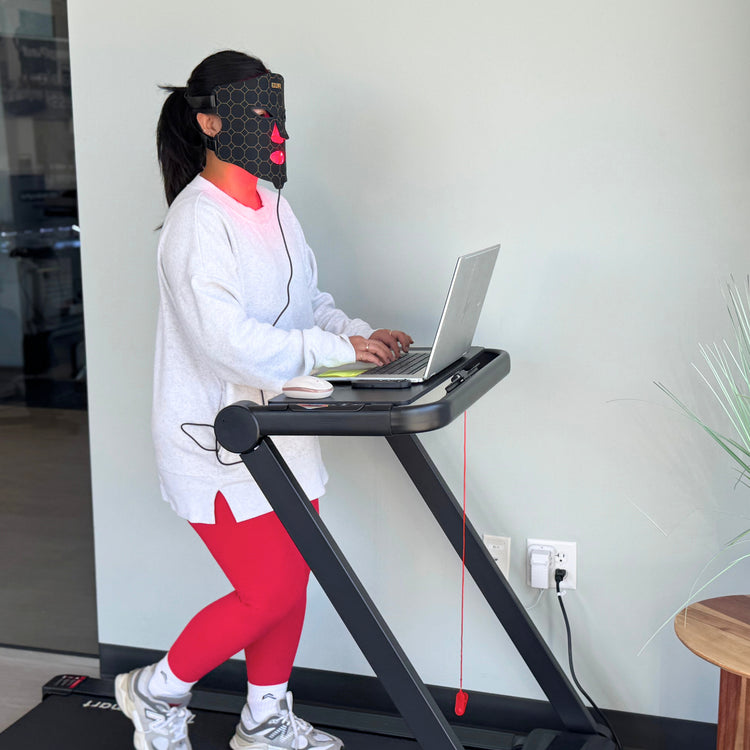Woman using a walking pad treadmill desk with a laptop, wearing a Red Light Mask with Infrared Light and vibration .