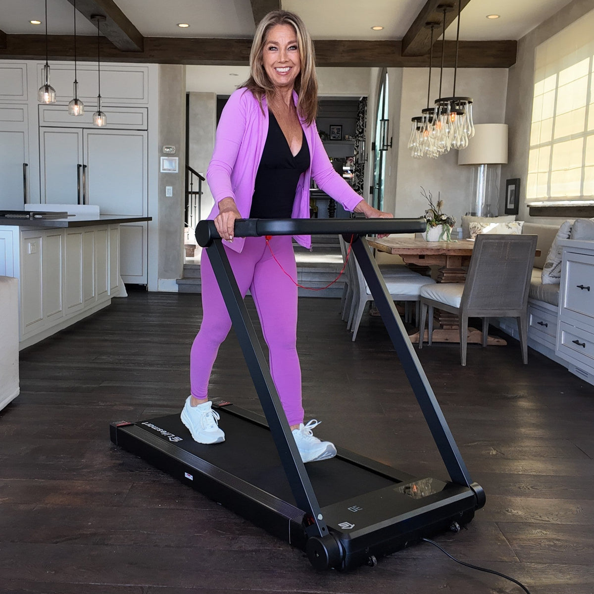 Denise Austin walking on her black treadmill in a modern kitchen.