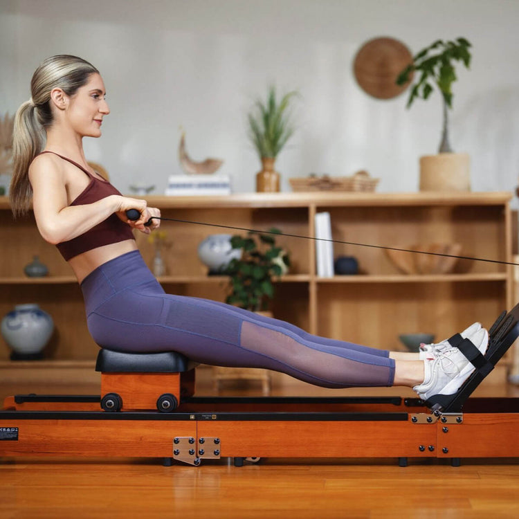 Woman using a foldable water rowing machine in a home setting, she's in the finish position with legs extended and arms pulled in.