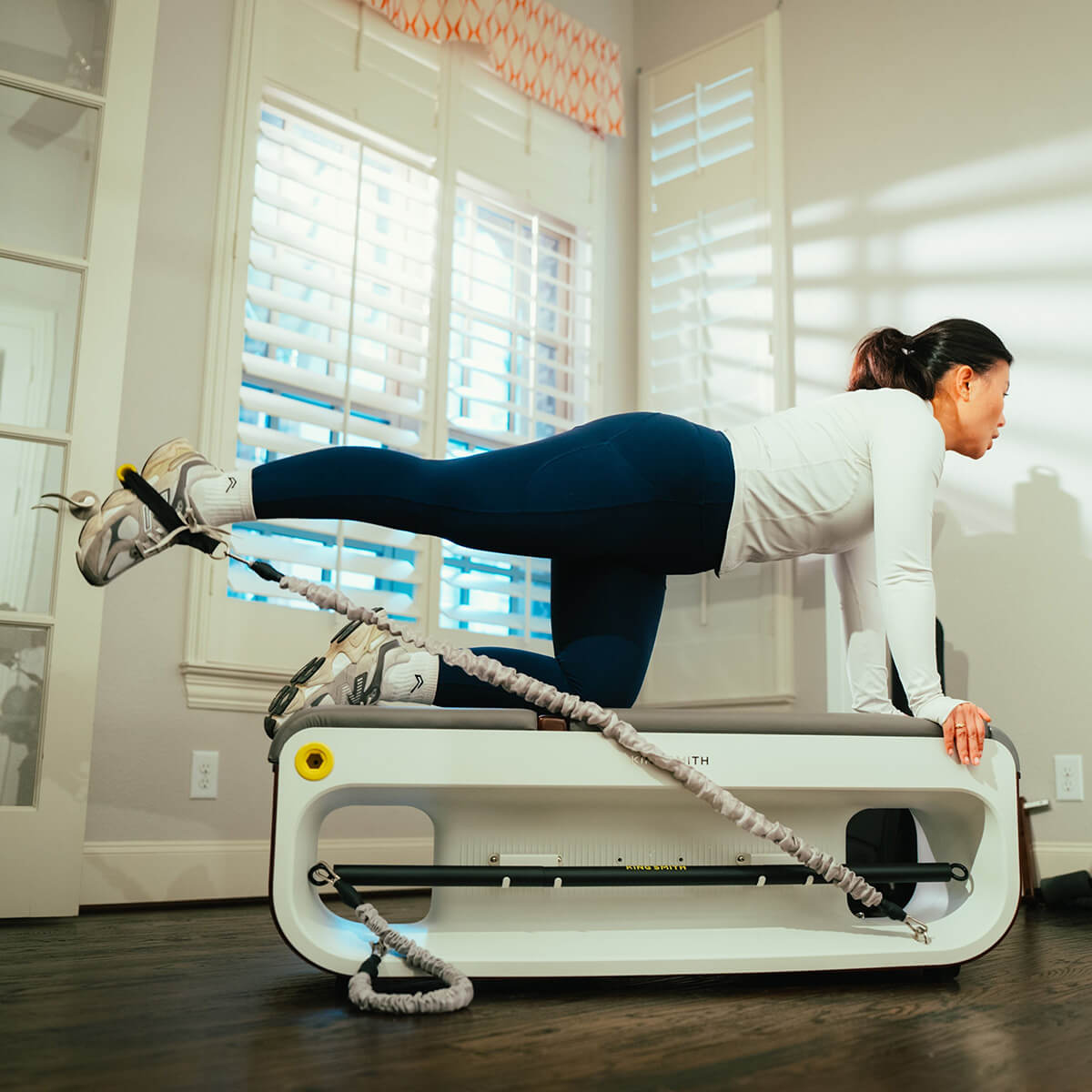 A person using a white and gray workout station bench for an exercise routine, with the equipment positioned in an indoor setting with wood flooring and large windows in the background.