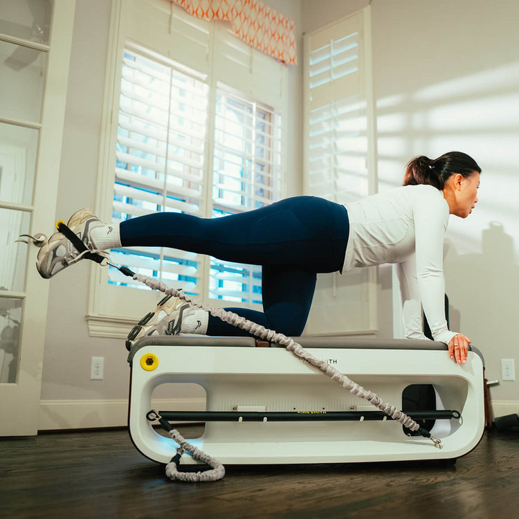 A person using a white and gray workout station bench for an exercise routine, with the equipment positioned in an indoor setting with wood flooring and large windows in the background.