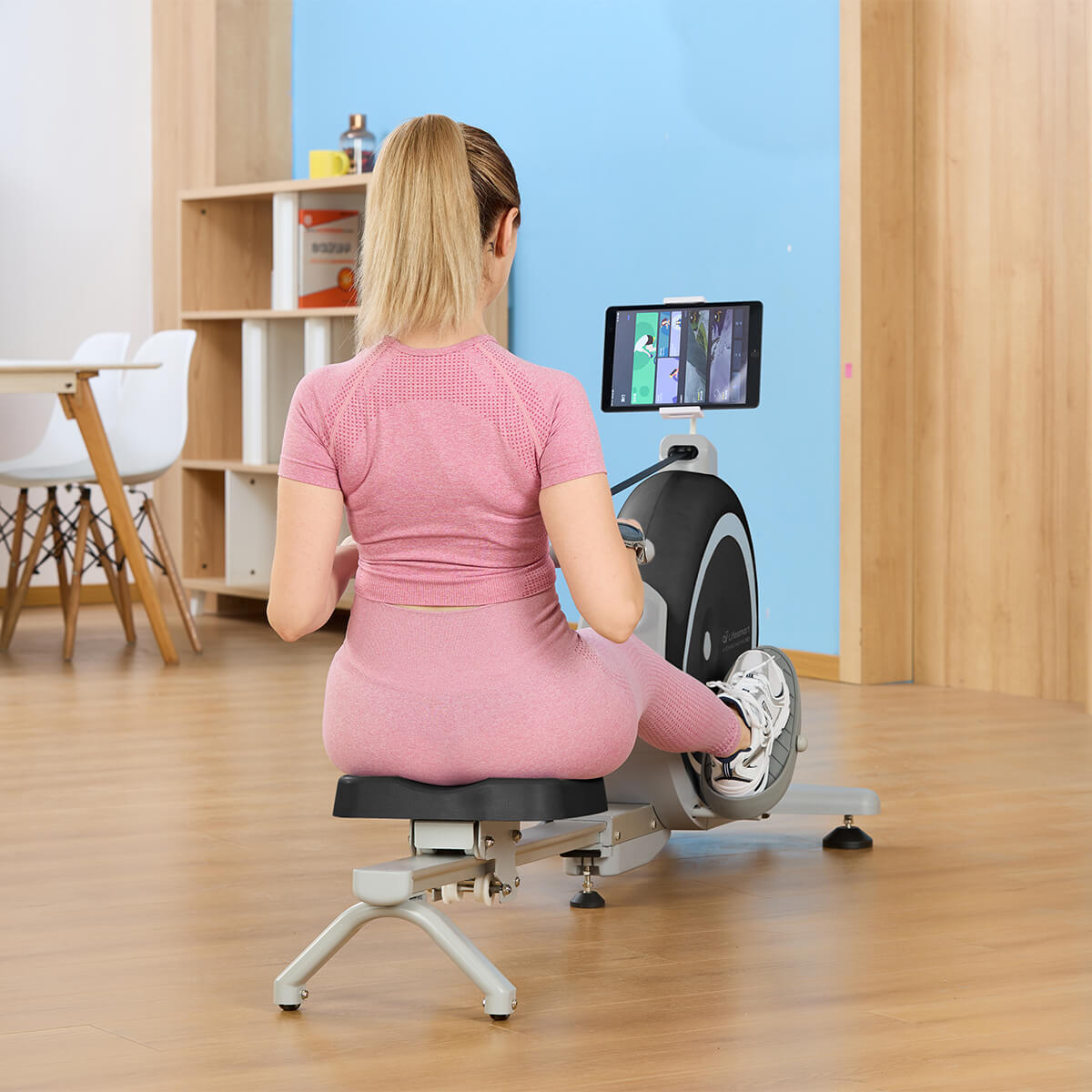 Woman in pink workout outfit exercising on rower. View from behind with woman looking at tablet in tablet holder.
